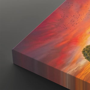 A close-up of a rectangular canvas print showing a dramatic sunset sky with red, orange, and pink clouds, small birds, and part of a tree, photographed on a dark gray background.