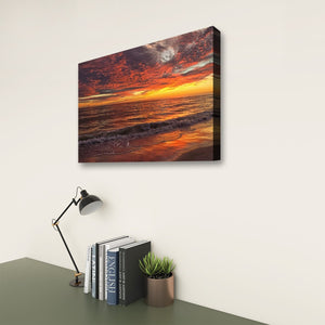 A rectangular wall art print showing a dramatic ocean sunset with orange, red, and dark clouds, displayed above a desk with a lamp, books, and a small plant against a light wall.