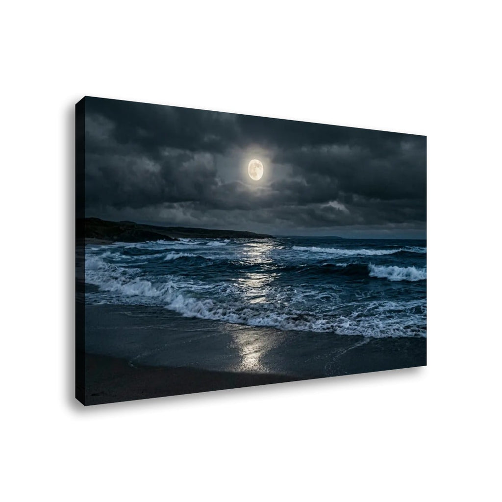A rectangular canvas wall art print showing a dramatic seascape at night under a full moon, with dark storm clouds, blue-gray waves, and wet sand reflecting light. The canvas is photographed at a slight angle against a plain white background.