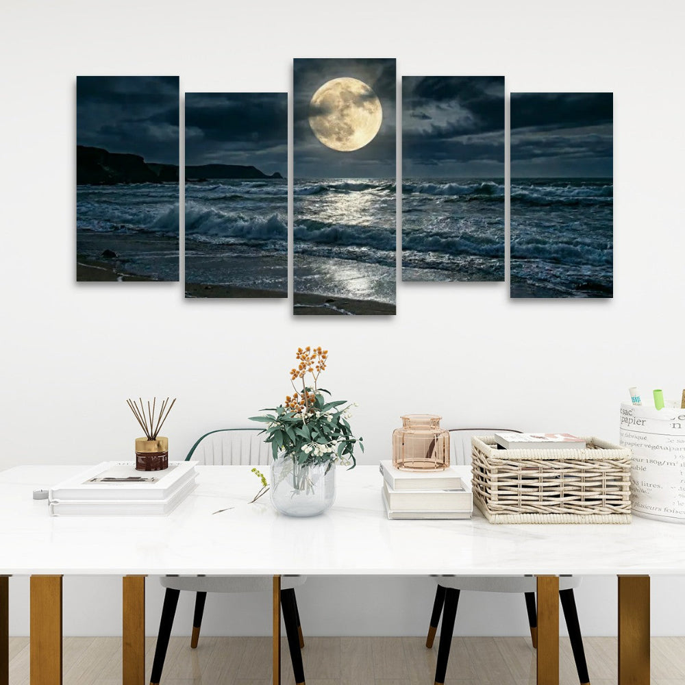 A 5-panel canvas wall art set depicting a dramatic night ocean scene with a large full moon, displayed above a white dining table in a modern room setting.