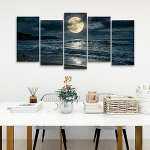 A 5-panel canvas wall art set depicting a dramatic night ocean scene with a large full moon, displayed above a white dining table in a modern room setting.