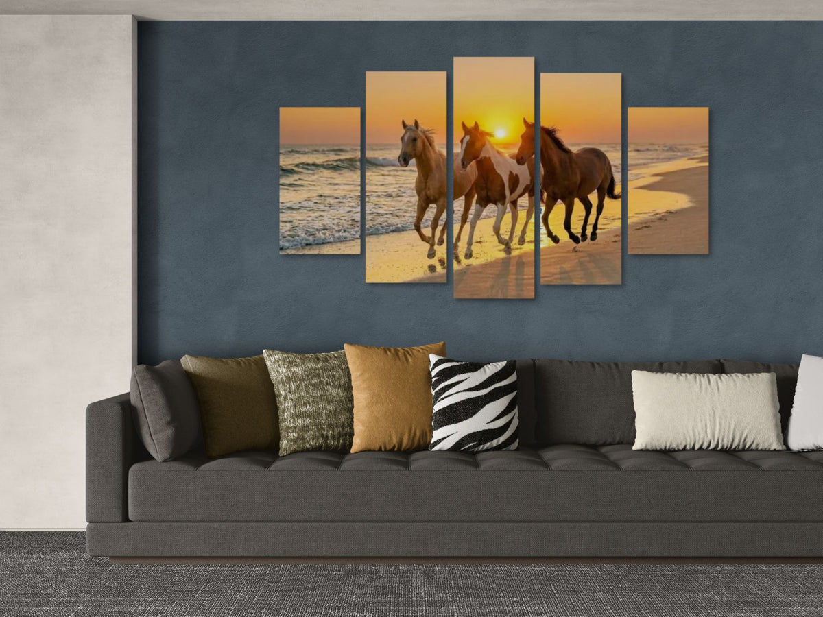 Five framed pictures of horses on a beach at sunset above a gray sofa with patterned cushions.