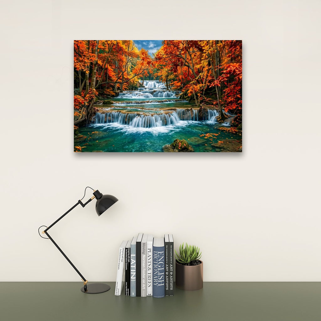 A rectangular wall art print showing a colorful autumn forest with a waterfall and turquoise water, displayed above a desk with a black lamp, books, and a small succulent on a light beige wall.