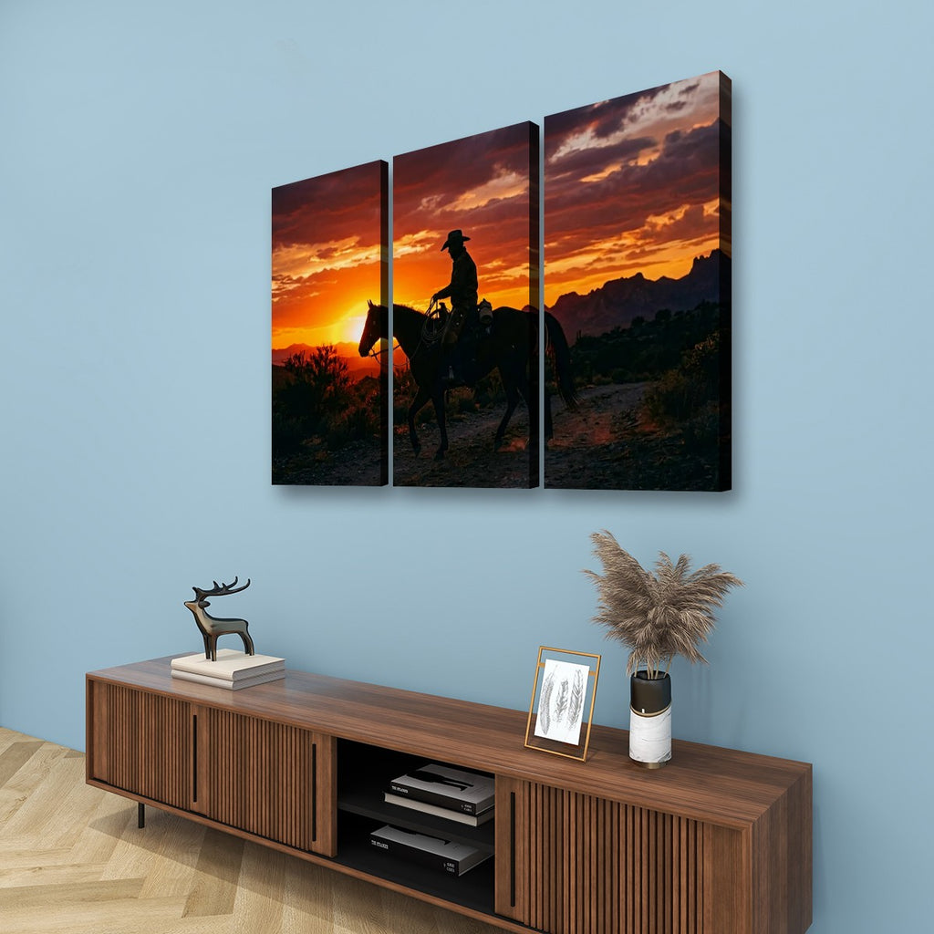 Three-panel canvas wall art showing a silhouetted cowboy riding a horse at sunset, displayed above a wooden media console against a light blue wall.