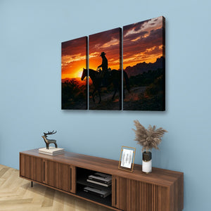 Three-panel canvas wall art showing a silhouetted cowboy riding a horse at sunset, displayed above a wooden media console against a light blue wall.