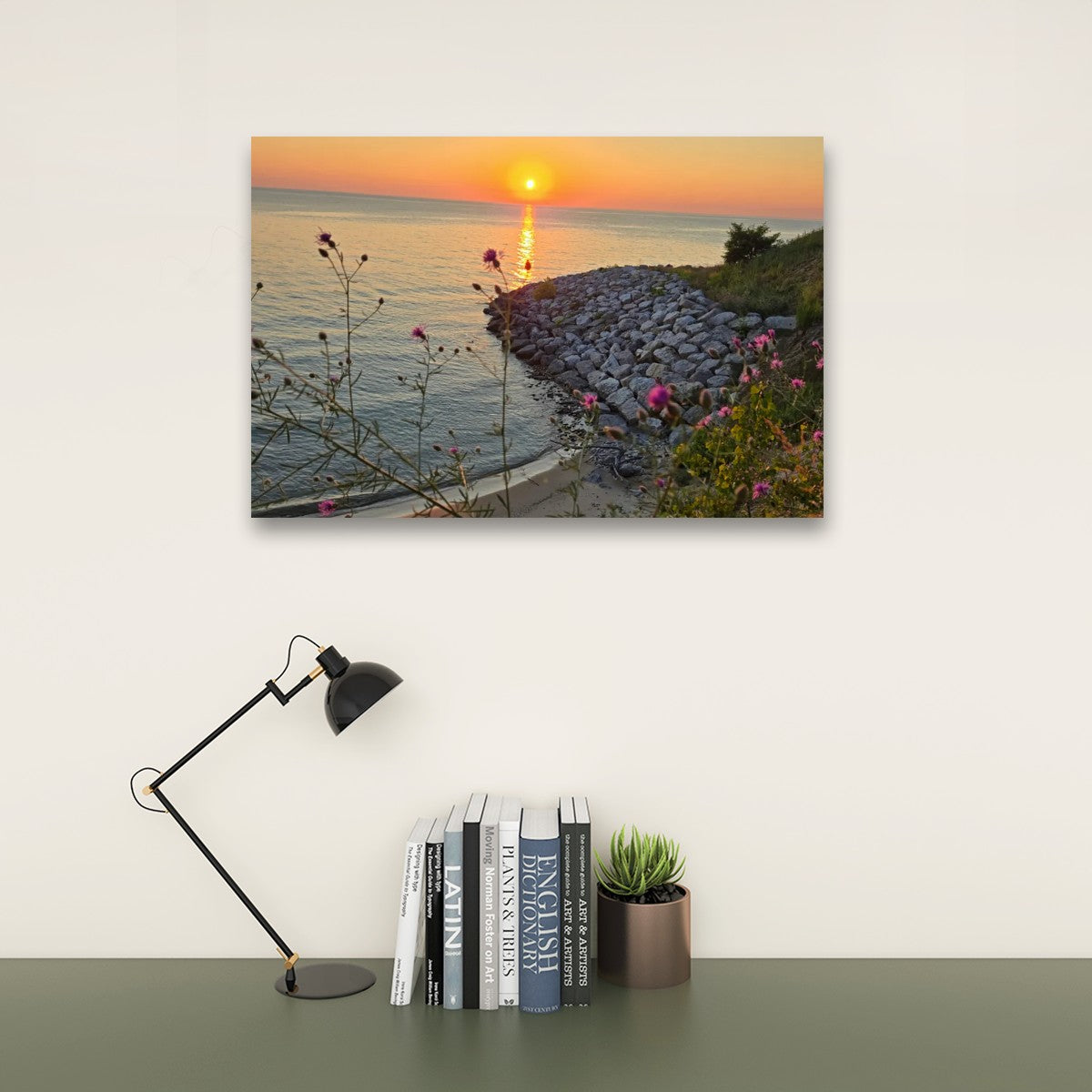 A rectangular wall art print showing a sunset over the water with a rocky shoreline and small pink flowers, displayed above a desk with a black lamp, books, and a small succulent against a light wall.