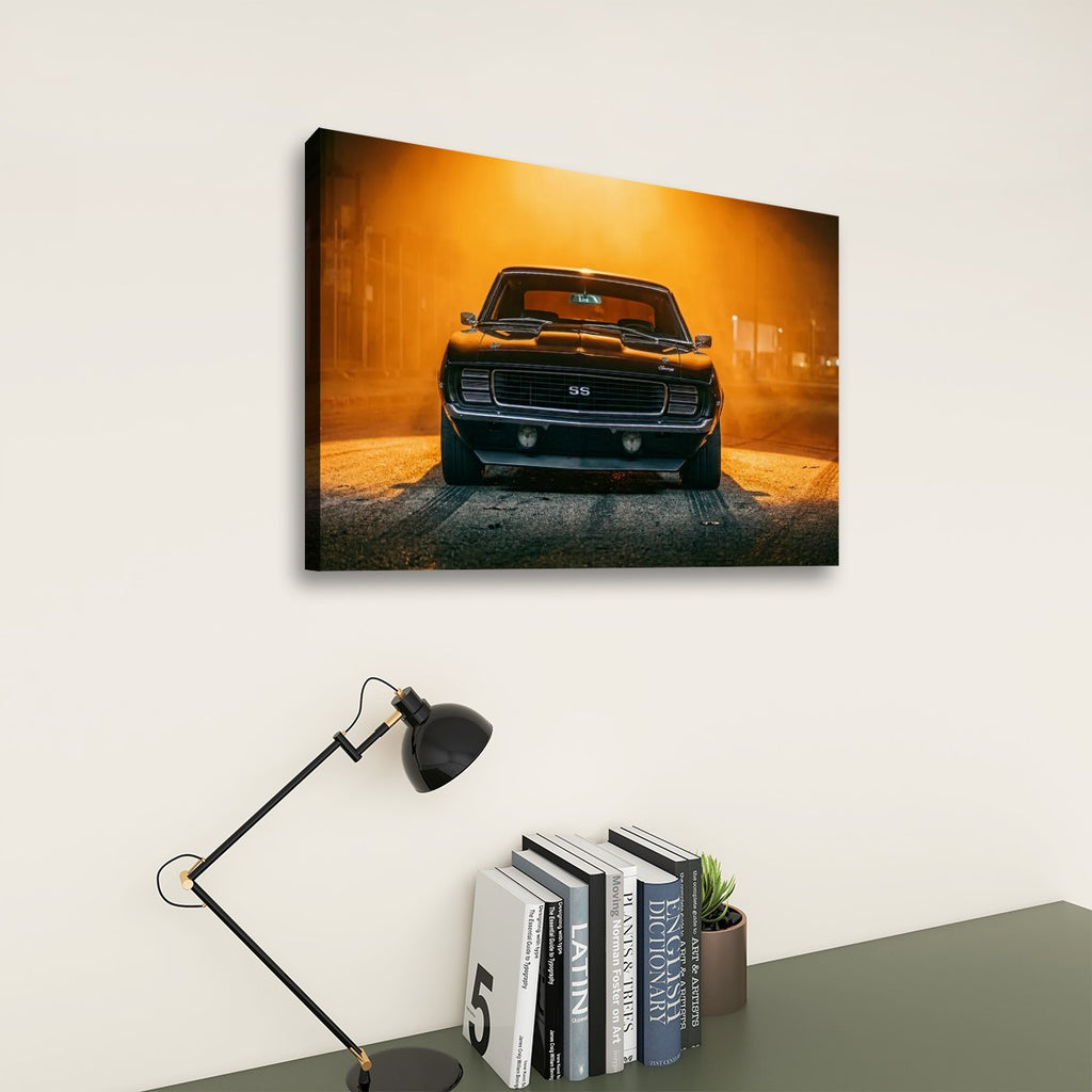 A rectangular wall art print showing a black classic muscle car in a warm orange-lit scene, displayed above a desk with a black lamp and books against a light beige wall.