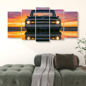A multi-panel canvas wall art print showing a black 1970 Chevy C10 facing forward on a reflective road at sunset, displayed above a green sofa in a living room setting.