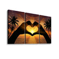 Three-panel canvas wall art showing a sunset seascape with palm trees and two silhouetted hands forming a heart shape over the ocean.