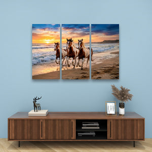 A three-panel canvas wall art print depicting three horses running on a beach at sunset, displayed on a light blue wall above a wooden console table with decorative items.