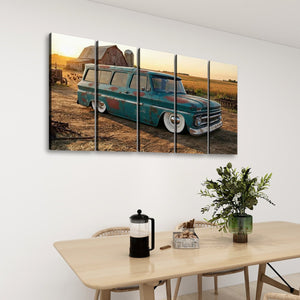 A five-panel canvas wall art print depicting a vintage teal 1966 Chevy Suburban Carryall station wagon parked in a rural field at sunset, displayed on a white wall above a dining table with chairs, a plant, and a coffee press.