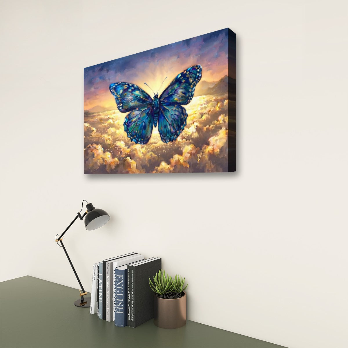 A rectangular wall art canvas showing a large blue butterfly over glowing clouds, displayed on a light wall above a desk with a lamp, books, and a small plant.