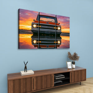 A three-panel canvas wall art print depicting a retro orange Ford truck reflected in water at sunset, displayed on a light blue wall above a wooden console table with decor.