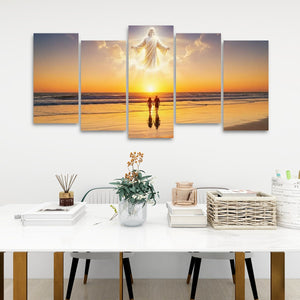 A five-panel canvas wall art set depicting a glowing figure standing over two small silhouetted figures at a beach sunset, displayed above a white table in a modern room setting.