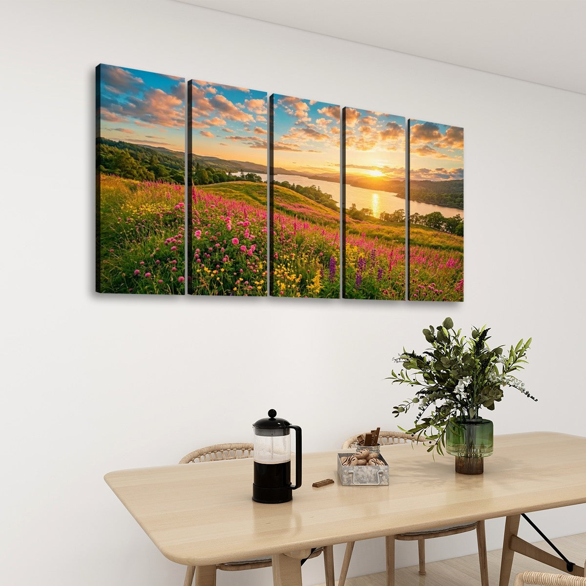 A five-panel canvas wall art set depicting a colorful sunset over a wildflower meadow and lake, displayed on a white wall above a dining table with a plant and coffee press.
