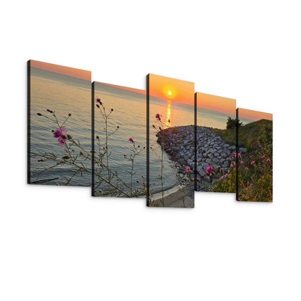 A five-panel canvas wall art set depicting a coastal sunset scene with calm water, a rocky shoreline, and pink wildflowers in the foreground, shown against a plain white background.