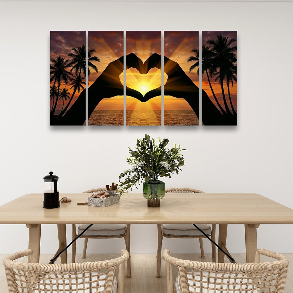 A five-panel canvas wall art set depicting a sunset over the ocean with palm trees and a heart-shaped hand gesture in the center, displayed on a white wall above a dining table with chairs and a vase.