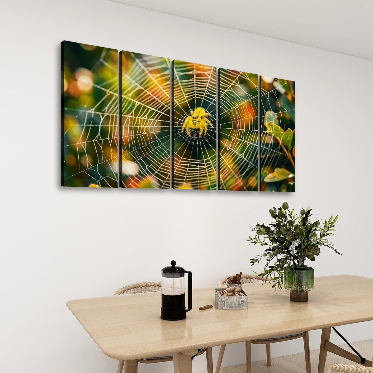 A five-panel canvas wall art set depicting a yellow spider on a detailed cobweb, displayed on a white wall above a light wood dining table with a vase of greenery and a French press.