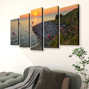 A five-panel canvas wall art set depicting a coastal sunset scene with rocks, flowers, and water, displayed on a white wall above a green sofa in a living room setting.