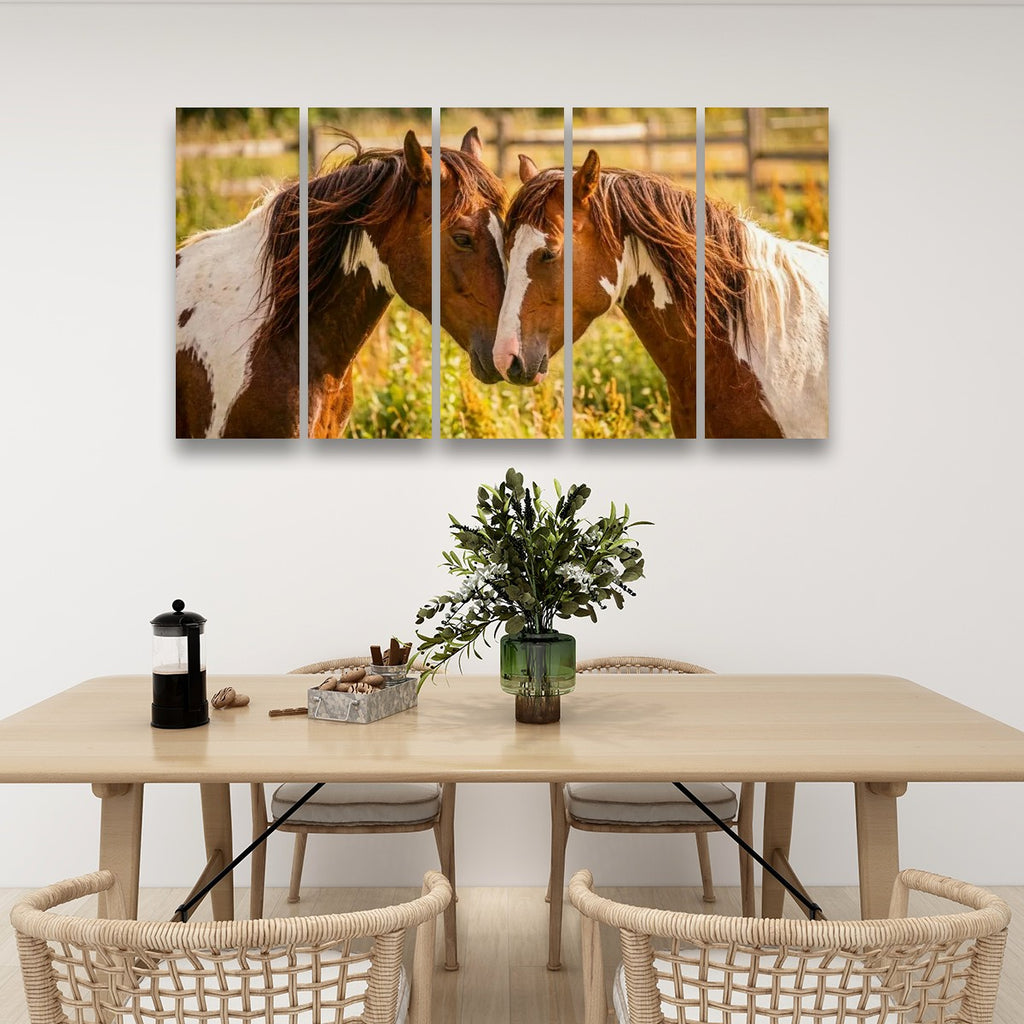 A five-panel canvas wall art set depicting two brown and white horses facing each other in a sunlit field, displayed above a light wood dining table with woven chairs in a modern dining room setting.