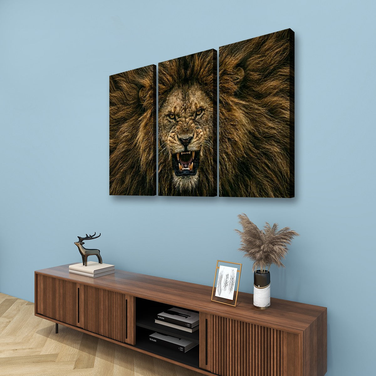 A three-panel canvas wall art set depicting a roaring lion's face and mane, displayed on a light blue wall above a wooden media console with decorative items.