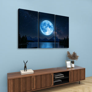 A three-panel canvas wall art set depicting a large glowing blue moon over mountains and a reflective lake at night, displayed above a wooden console in a styled room.