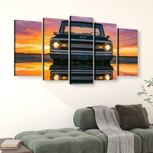 A five-panel canvas wall art set depicting a front view of an orange 1970 Chevy C10 truck reflected on water at sunset, displayed above a green sofa in a living room setting.