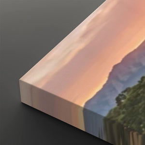A close-up of a rectangular canvas or print showing a landscape scene with a sunset sky in peach and pink tones, mountains, and dark green trees, photographed on a dark gray background.