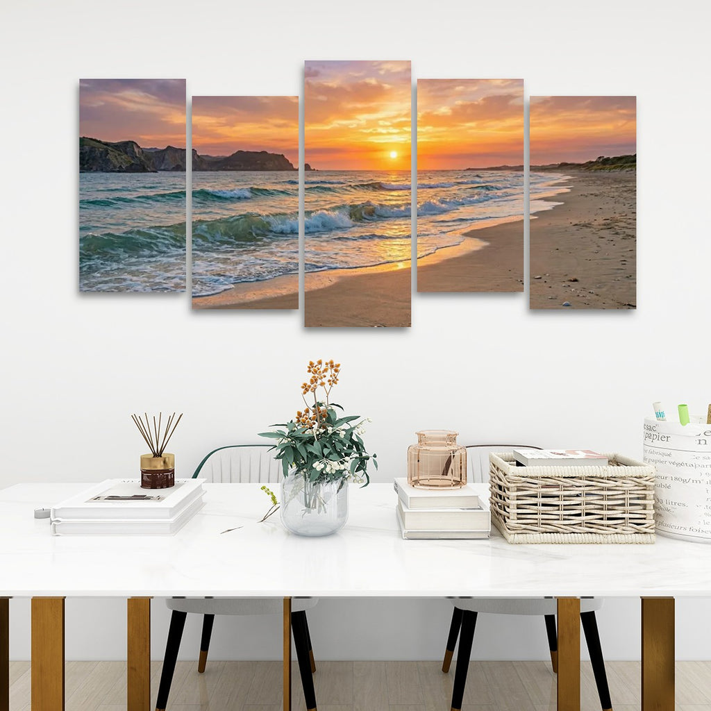 A five-panel canvas wall art set depicting a beach sunset with ocean waves, displayed above a white dining table in a modern room setting.