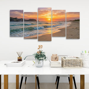 A five-panel canvas wall art set depicting a beach sunset with ocean waves, displayed above a white dining table in a modern room setting.