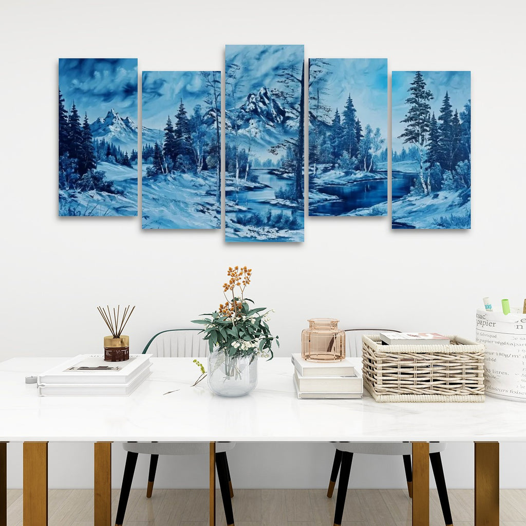 A five-panel canvas wall art set depicting a snowy mountain landscape in shades of blue, displayed on a white wall above a styled dining table with books, a vase, and a basket.