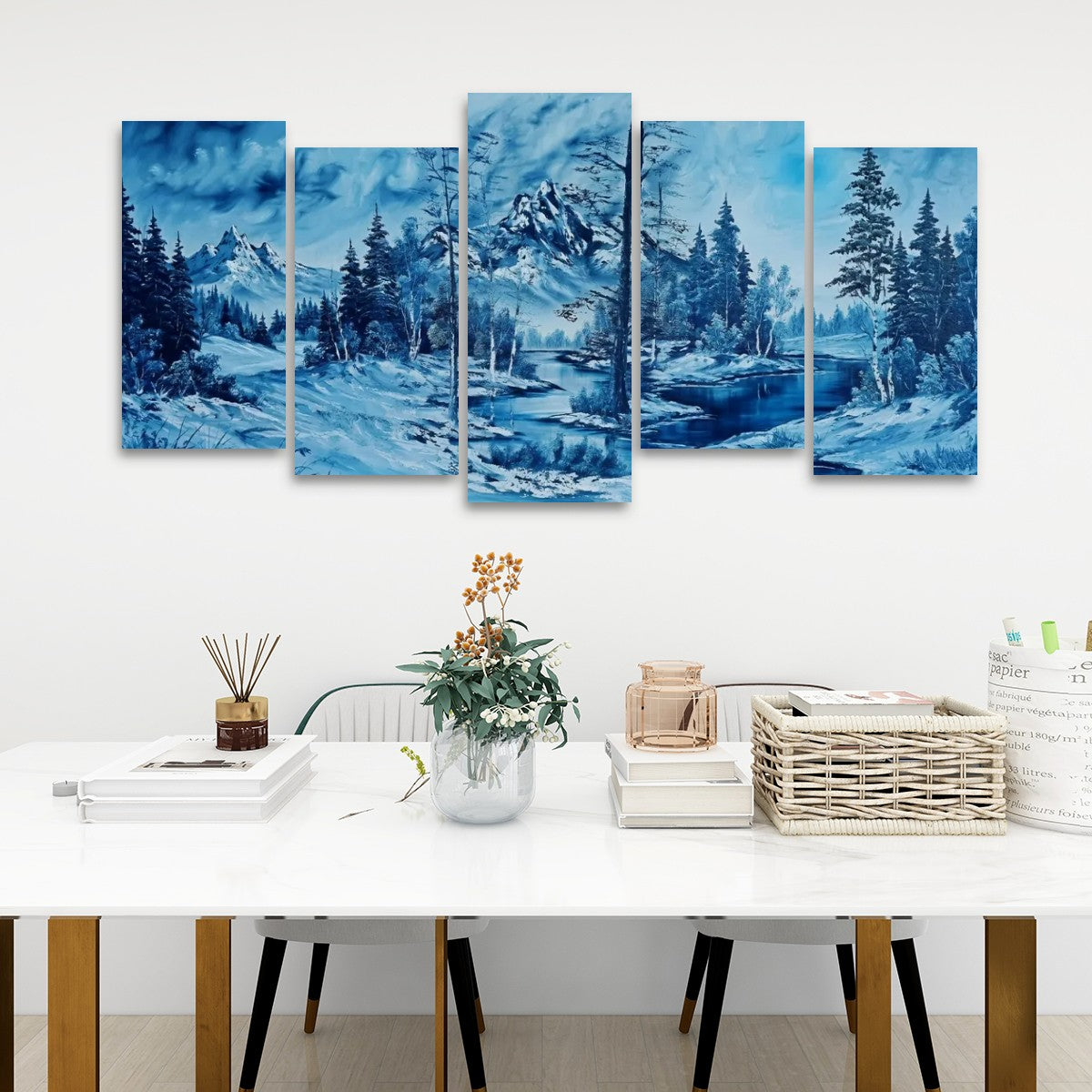 A five-panel canvas wall art set depicting a snowy mountain landscape in shades of blue, displayed on a white wall above a styled dining table with books, a vase, and a basket.