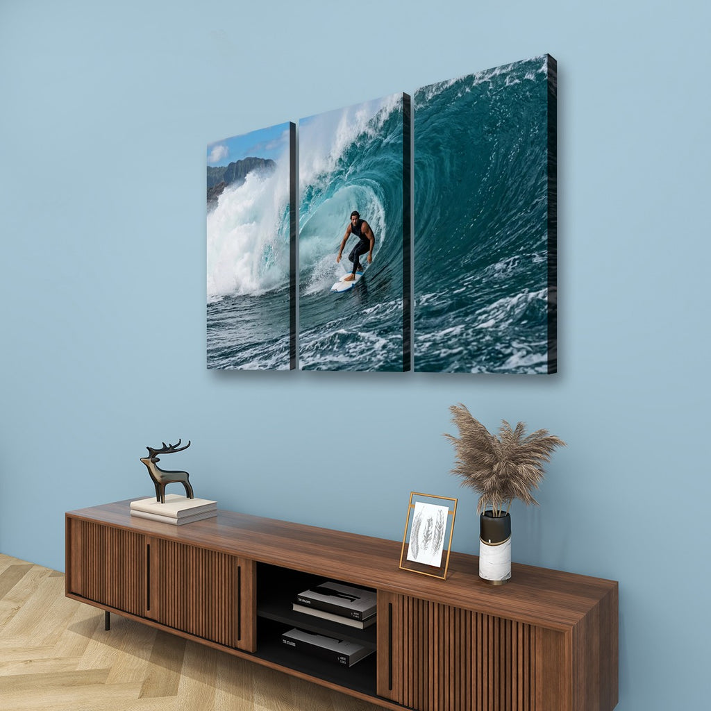 A three-panel canvas wall art set depicting a surfer inside a large curling ocean wave, displayed on a light blue wall above a wooden console table with decorative items.