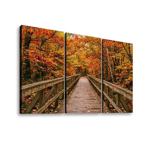 A three-panel canvas wall art print depicting a wooden boardwalk bridge spanning through an autumn forest with vibrant orange, red, and yellow foliage.