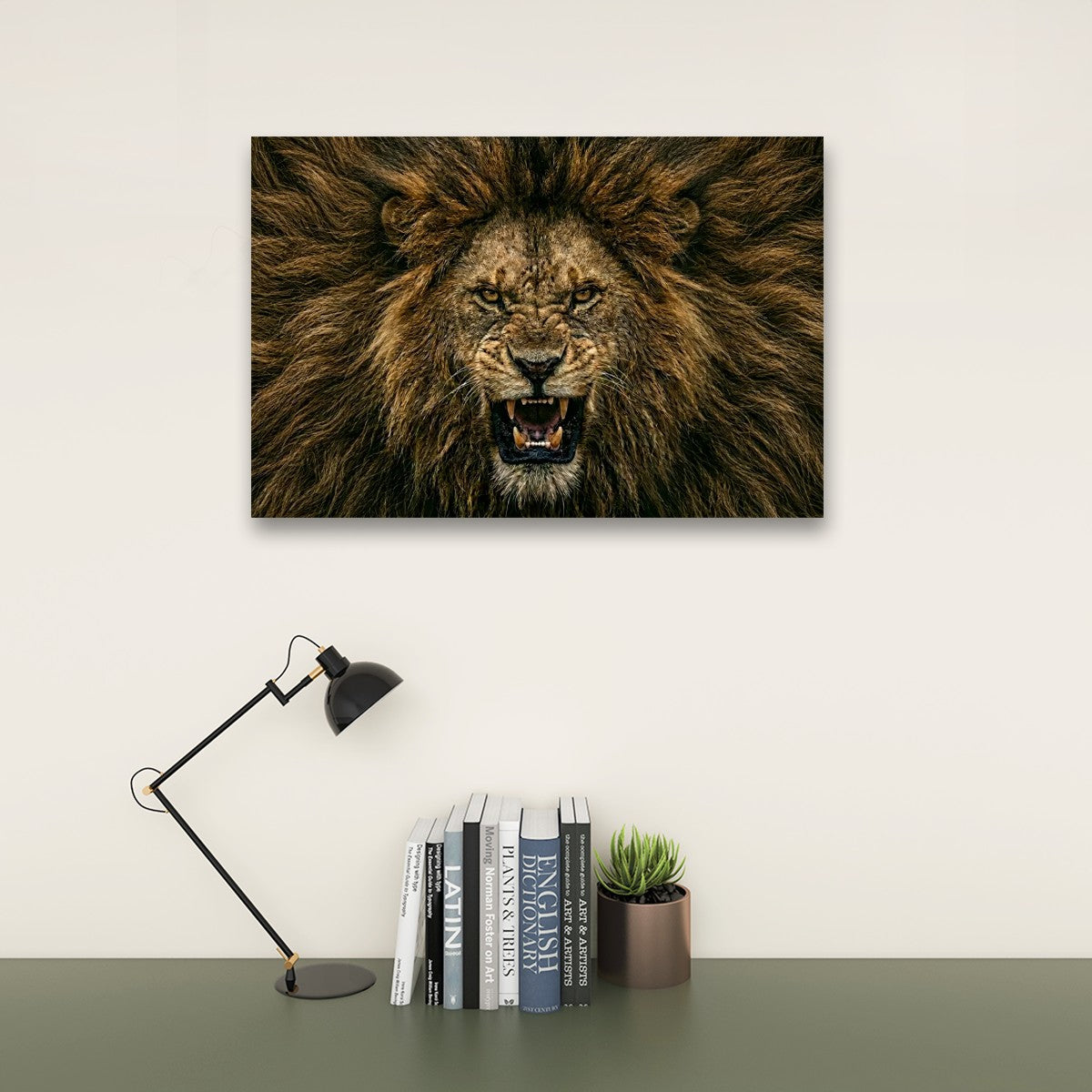 A rectangular wall art print showing a close-up lion face with an open mouth, displayed above a desk with a black lamp, books, and a small succulent against a light wall.
