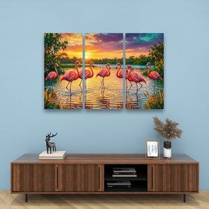 A three-panel canvas wall art set depicting pink flamingos standing in a lake at sunset, displayed on a light blue wall above a wooden console table with decorative items.