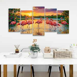 A five-panel canvas wall art set depicting pink flamingos standing in a lake at sunset, displayed on a white wall above a modern white table with decorative items.