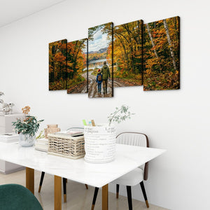 A five-panel canvas wall art set depicting an autumn forest path with two people walking, displayed on a white wall above a dining table in a modern room setting.