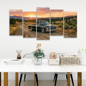 A five-panel canvas wall art set depicting a vintage pickup truck on a dirt road at sunset, displayed on a white wall above a styled dining table with books, a vase, and chairs.