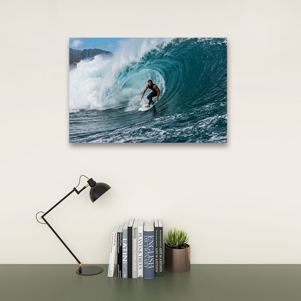A wall-mounted photographic print of a surfer riding a large blue ocean wave, displayed above a desk with a black lamp, books, and a small potted succulent against a light beige wall.