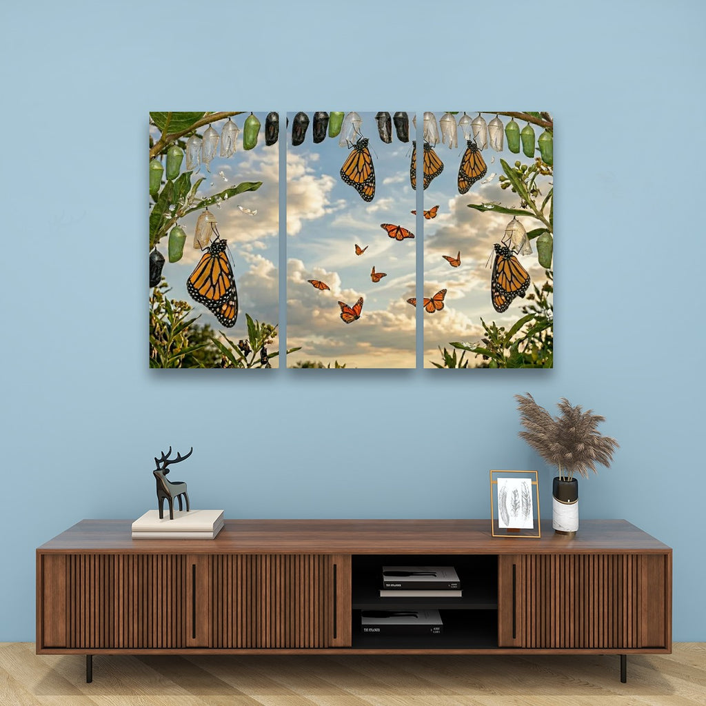 A three-panel canvas wall art print depicting monarch butterflies and chrysalises hanging from a tree against a cloudy sky, displayed on a light blue wall above a wooden console table.