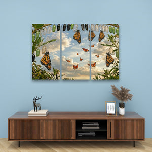 A three-panel canvas wall art print depicting monarch butterflies and chrysalises hanging from a tree against a cloudy sky, displayed on a light blue wall above a wooden console table.
