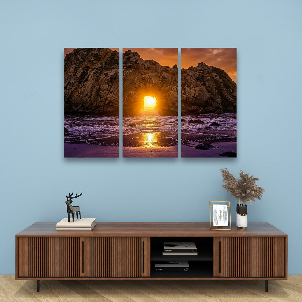 A three-panel canvas wall art set depicting a dramatic rocky coastline at sunset, hung on a light blue wall above a wooden console table with decorative items.