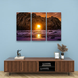 A three-panel canvas wall art set depicting a dramatic rocky coastline at sunset, hung on a light blue wall above a wooden console table with decorative items.