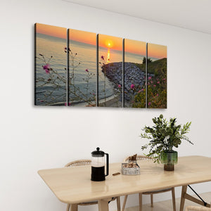 A five-panel canvas wall art set depicting a scenic sunset over a rocky shoreline with wildflowers, displayed on a white wall above a dining table with chairs, a coffee press, and a potted plant.