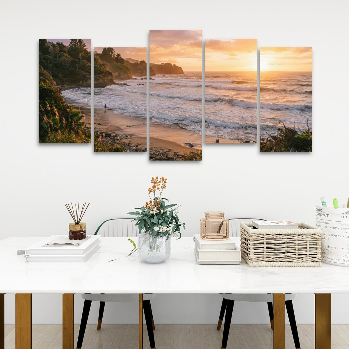 A five-panel canvas wall art set depicting a coastal beach sunset, displayed on a white wall above a styled dining table with books, a vase, and baskets.