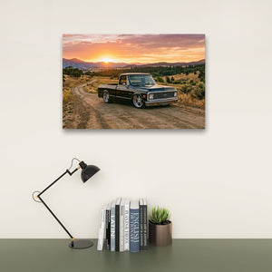 A rectangular wall art print showing a vintage 1970 Chevy C10 Fleetside pickup truck on a dirt road at sunset, displayed above a desk with a black lamp, books, and a small succulent against a light wall.