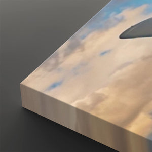 A close-up of a rectangular canvas or panel showing a printed sky scene with soft blue clouds and a dark aircraft wing silhouette, photographed on a dark gray background.