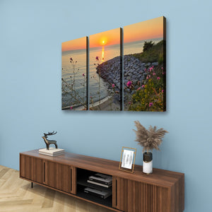 A three-panel canvas wall art set depicting a coastal sunset over water with a rocky shoreline and pink flowers, displayed on a light blue wall above a wooden console table.