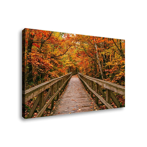 A rectangular canvas wall art print showing a wooden boardwalk bridge spanning through an autumn forest with orange, yellow, and red foliage, photographed against a plain white background.